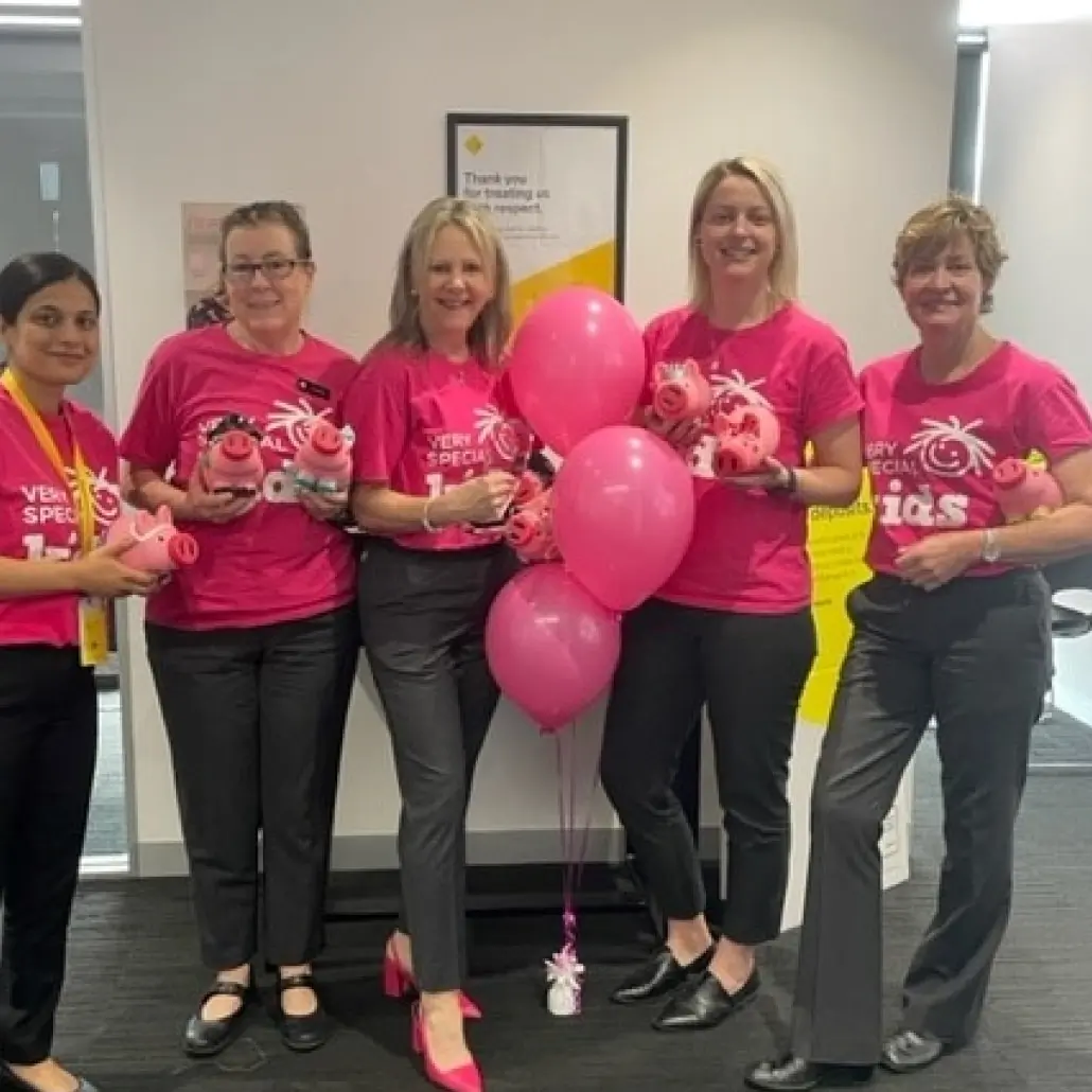 A group of adults are standing together holding piggy banks and wearing pink Very Special Kids t-shirts