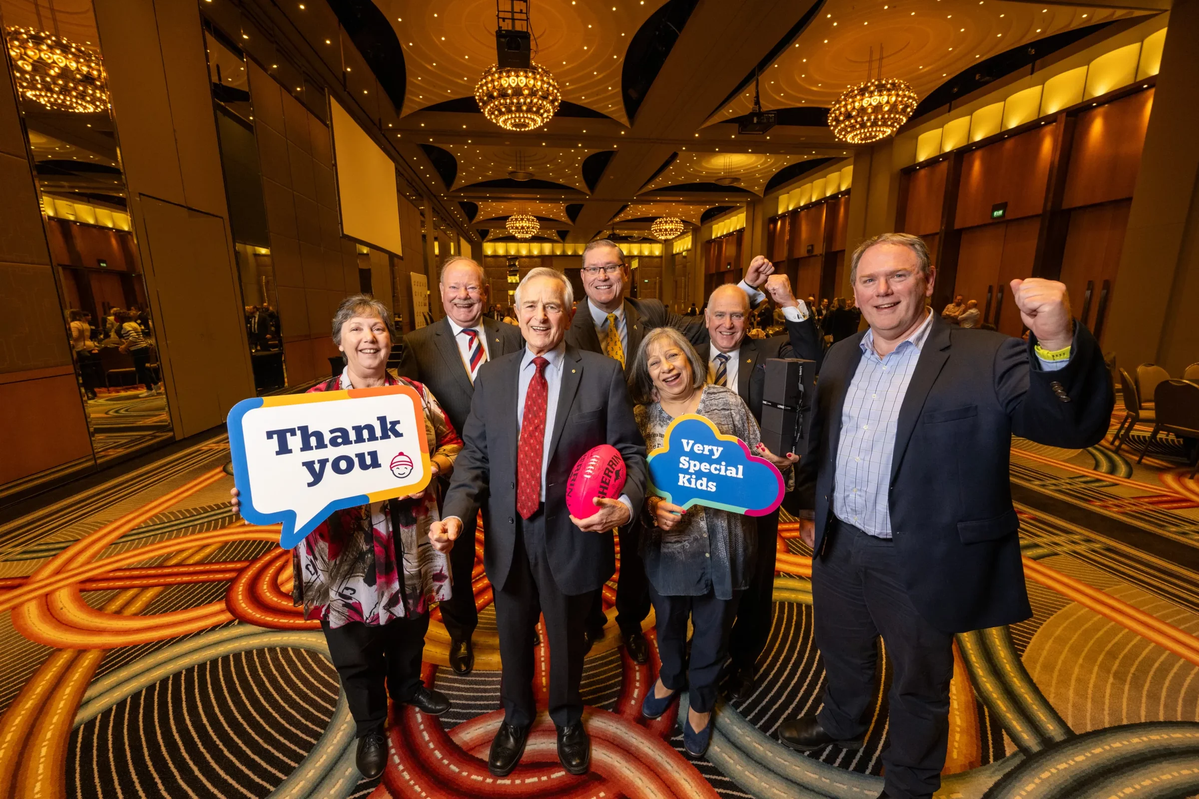 A group of adults are standing together in a function room. They are holding a football and signs reading "Thank you" and "Very Special Kids"