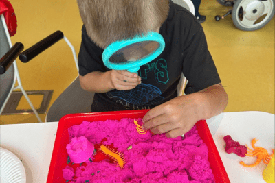 A boy is looking through a magnifying glass at colourful sand and toy bugs